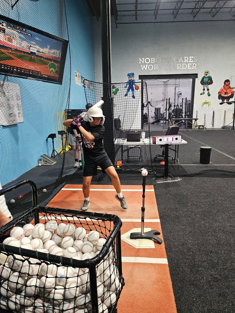 Person practicing batting in a indoor sports facility with a screen displaying a baseball game.