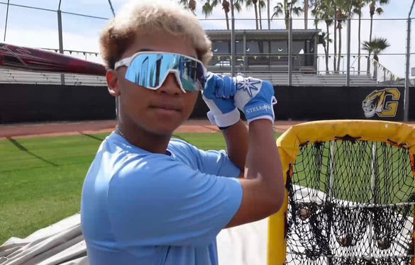 Person wearing blue sunglasses and gloves, standing on a sports field with a net in the background.