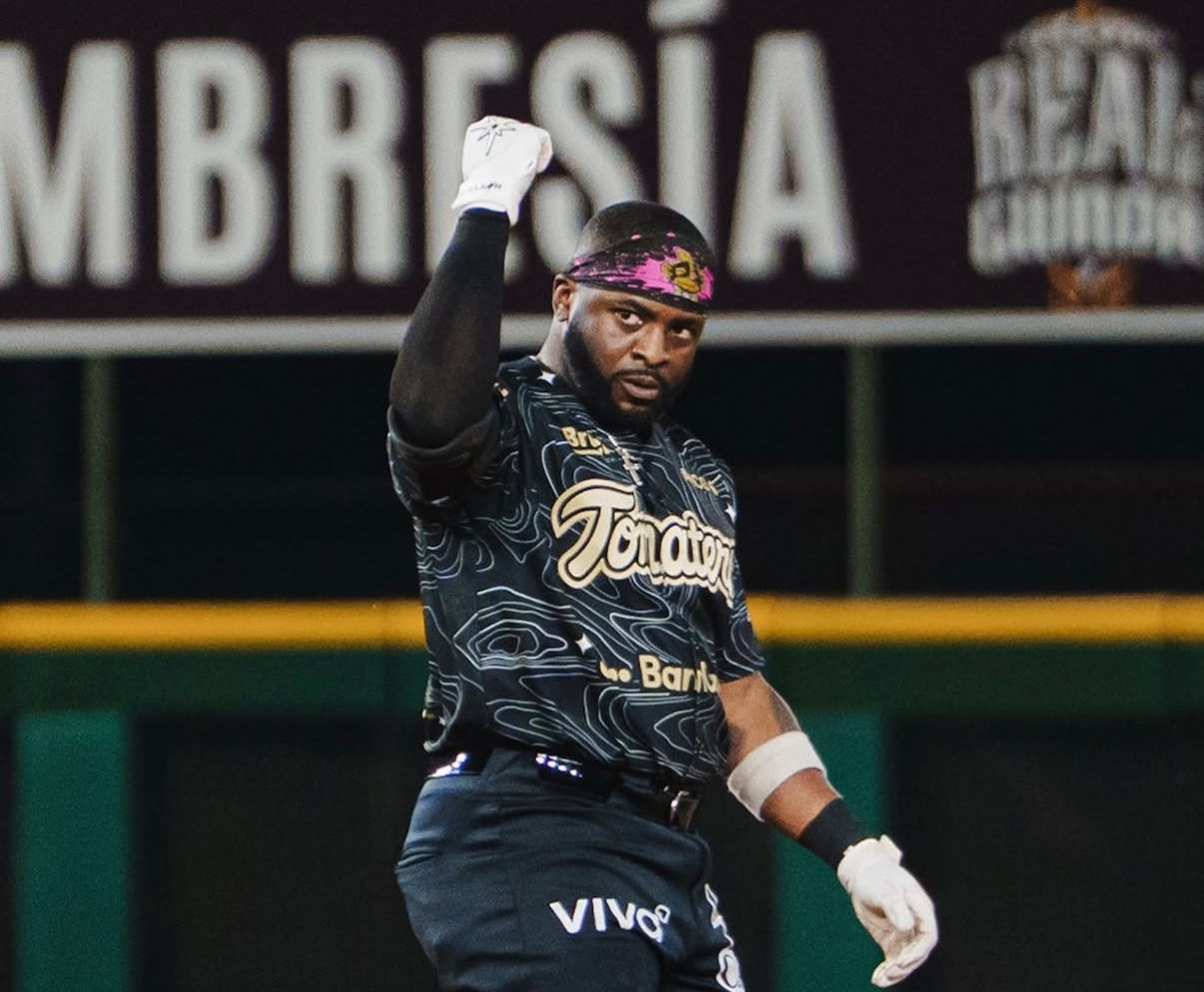 Baseball player in a dark uniform with logos, standing on a baseball field.