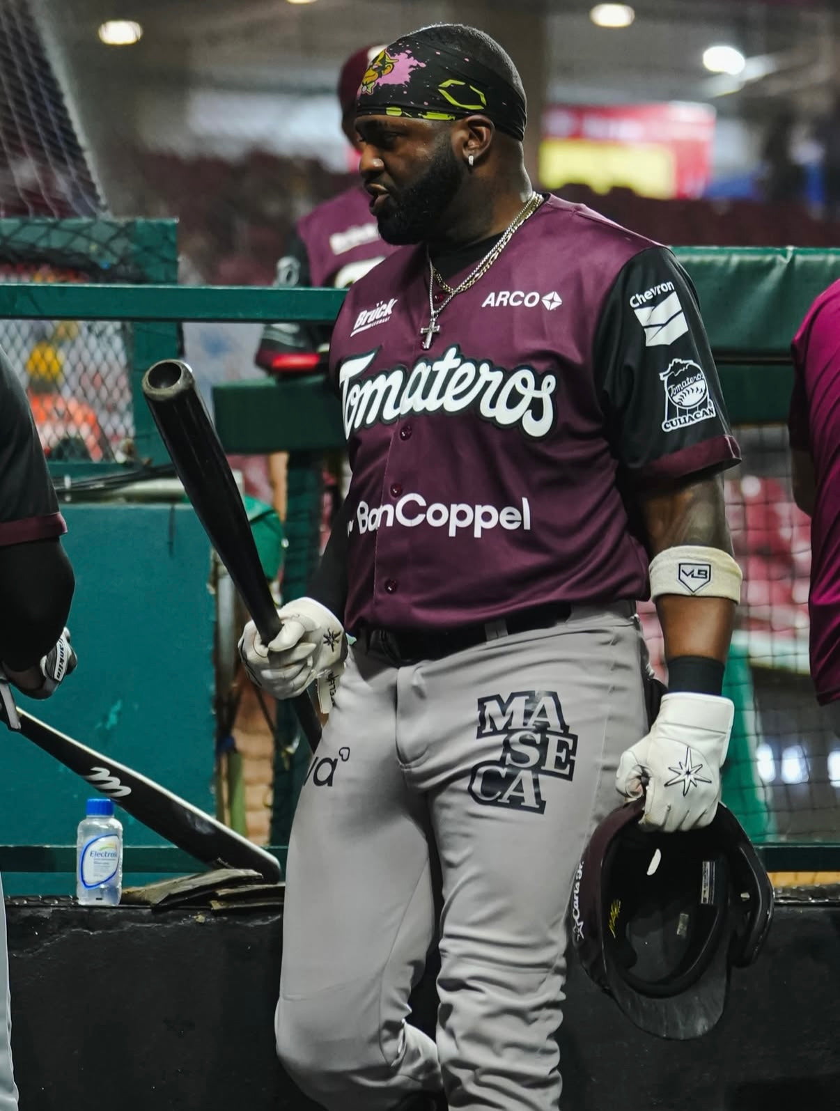 Baseball player in maroon and gray uniform with 'Tomateros' and 'Bon Coppel' branding, holding a bat and helmet.