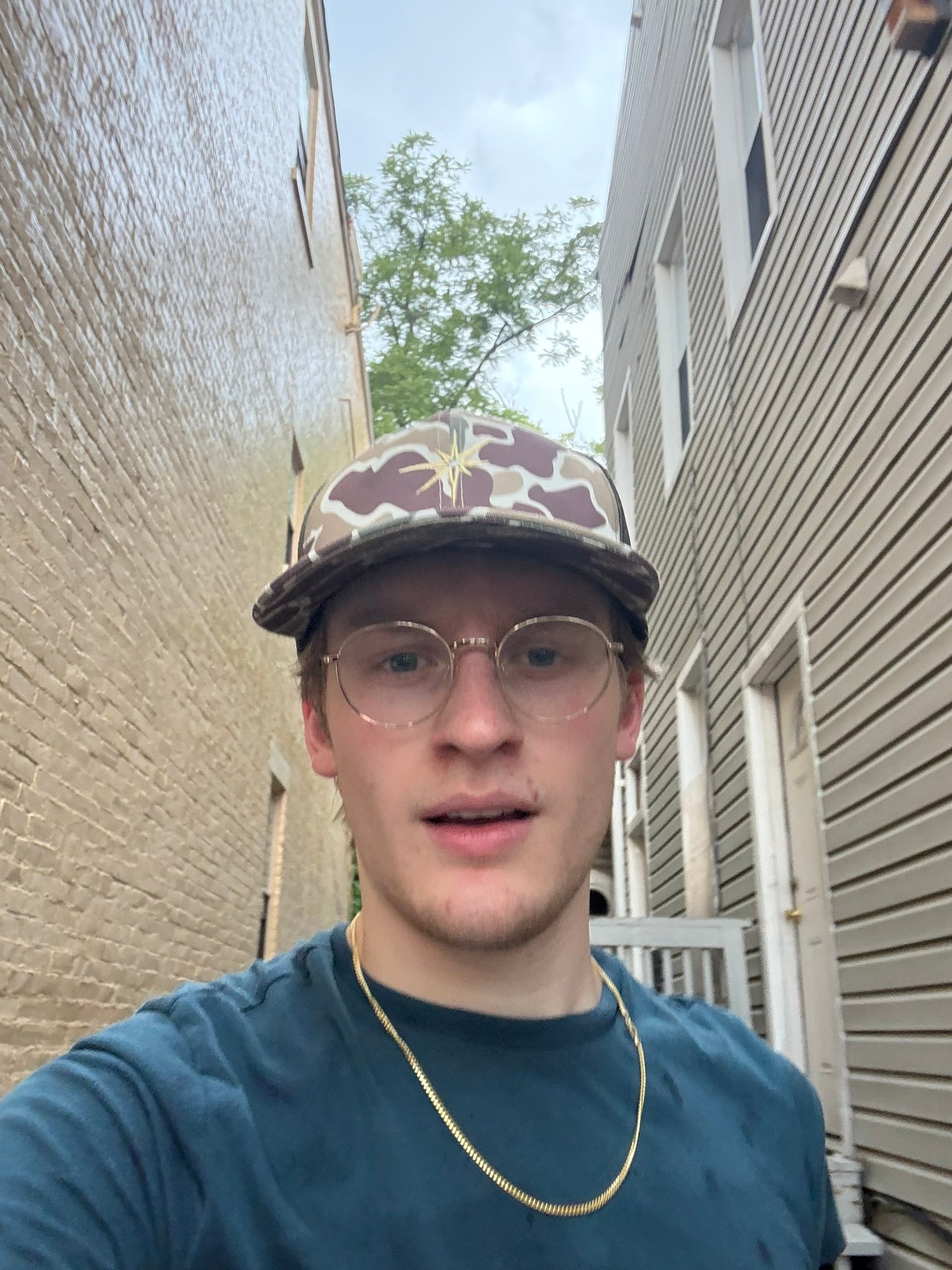 Person wearing a cap and glasses standing in an alleyway between buildings.